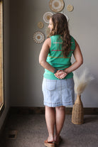 Woman in a green sleeveless top and denim skirt standing in a room with decorative wall art.