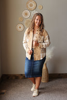 A woman modeling a beige and white plaid button-up shirt jacket over a cream top, paired with a blue denim skirt and beige sneakers, standing in a room with boho wall decor.