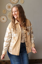A woman wearing a tan and beige plaid button-up shirt over a beige top and blue jean skirt, standing in front of a neutral wall with woven wall decor.