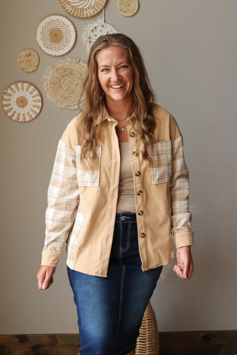 A woman modeling a tan button-up shirt jacket with beige and white plaid sleeves and chest pockets, worn over a beige top and blue jean skirt, standing against a neutral wall with woven wall decor.