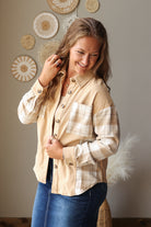 A woman wearing a beige and white plaid button-down shirt with blue jean skirt, standing in a room with decorative wall hangings.