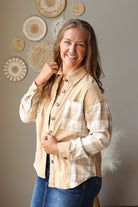 A woman smiling and posing in a beige and white plaid button-down shirt with blue jean skirt, standing against a neutral wall decorated with circular woven wall art.