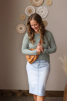 Woman wearing a green sweater and light blue skirt holding a brown bag against a wall with decorative items. Experience comfort and style with the Lagoon Blue Waffle Top, perfect for casual days and effortless layering.