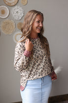 A woman wearing a beige leopard print long-sleeve top with red and white polka dot cuffs and hem, paired with light blue jean skirt, standing against a gray wall decorated with circular woven wall art.