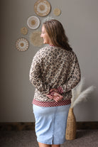A woman is shown from the back wearing a beige and brown leopard print long-sleeve top with a red and white polka dot hem, paired with a light blue denim skirt. She is standing in a room with a gray wall decorated with circular woven wall art.