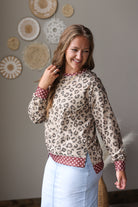 A woman wearing a beige leopard print long-sleeve top with red and white polka dot trim, paired with light blue jean skirt, standing against a neutral wall decorated with circular woven wall art.