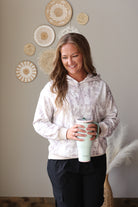 A woman smiling while holding a mint green travel tumbler, wearing a white and gray camo patterned hoodie and dark cargo pants, standing in front of a beige wall decorated with circular woven wall art.