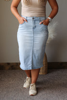 A person wearing a light blue denim midi skirt with a front fly and side pockets, paired with a beige top and white sneakers, standing indoors against a neutral wall.