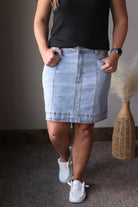 Person wearing a black top and light blue denim skirt with white sneakers, standing against a neutral background.