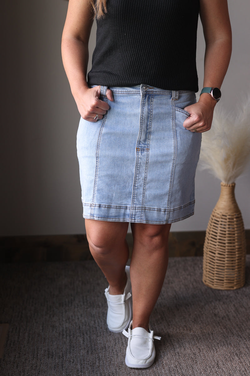Person wearing a black top and light blue denim skirt with white sneakers, standing against a neutral background.