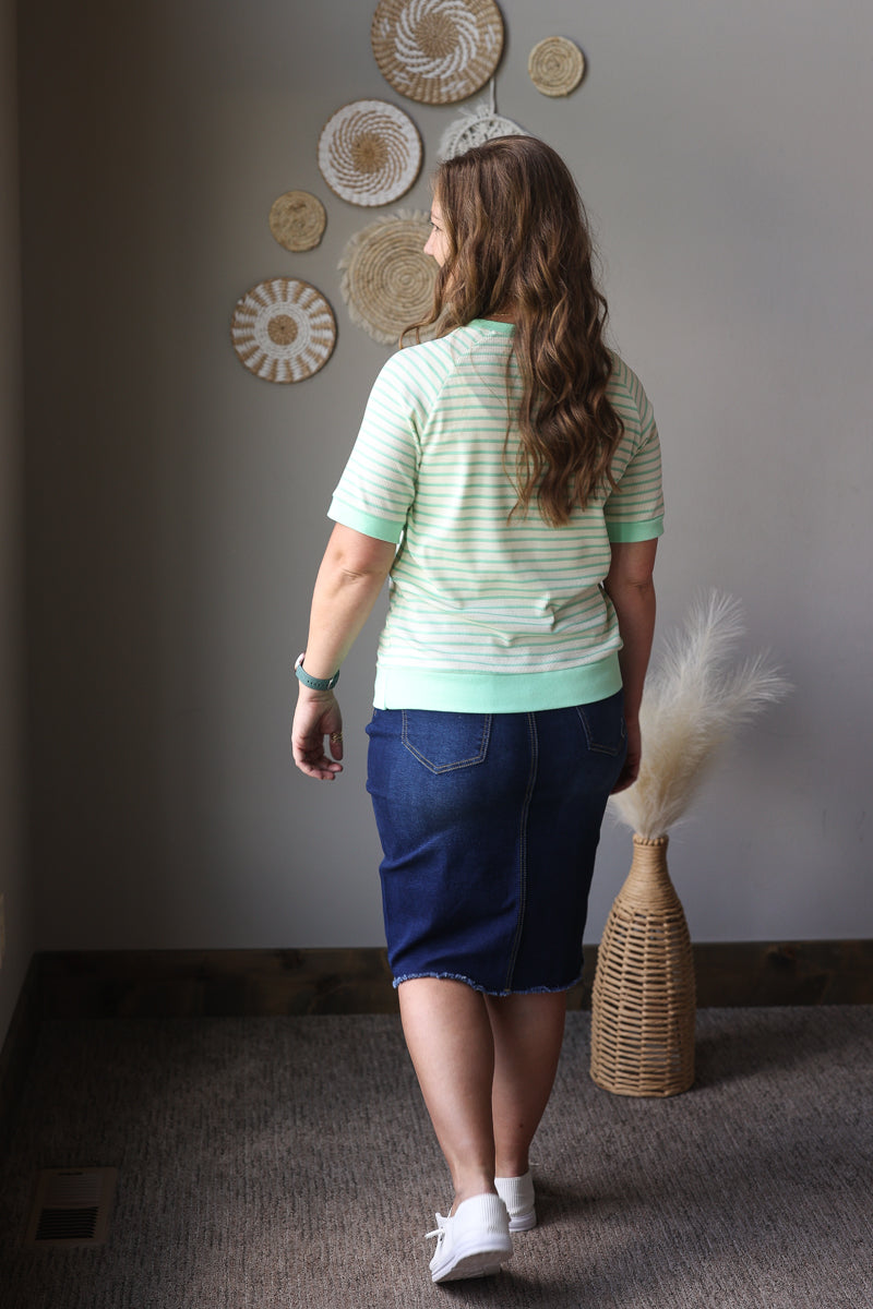Person wearing a green striped shirt and blue denim skirt standing in a room with decorative wall art.