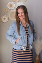 Woman wearing a denim jacket over a striped dress, standing against a wall with decorative items.