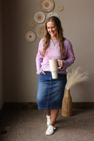 Woman in a purple spring top and blue skirt holding a white mug, standing in a room with decorative wall art.