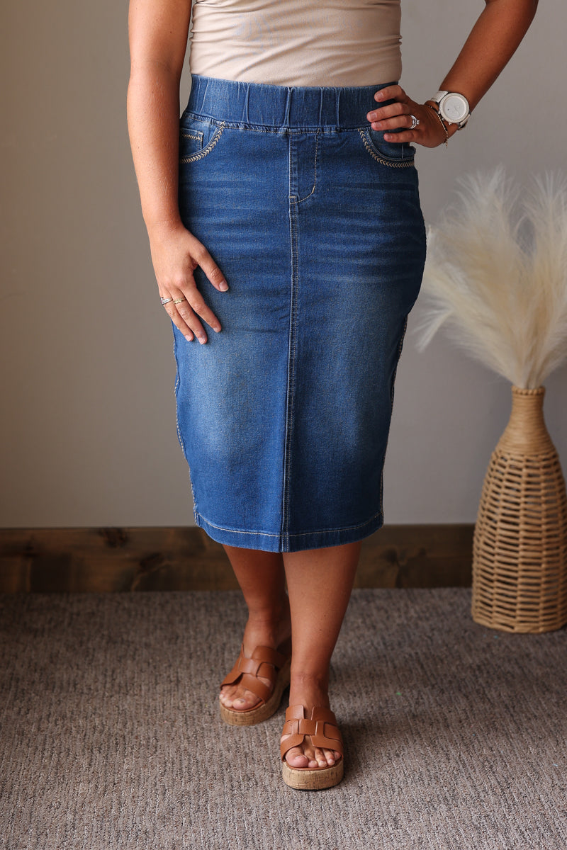 A woman wearing a mid-length blue denim skirt, paired with a beige top and brown platform sandals, standing in an indoor setting with a neutral wall and a decorative woven vase.