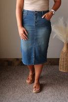 A person wearing a medium-wash blue denim midi skirt with front and back seam detailing, paired with tan leather platform sandals and a beige top, standing on a textured gray carpet.