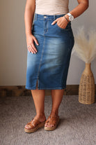 A person wearing a blue denim midi skirt with a front slit, paired with a beige top and brown espadrille sandals, standing indoors on a textured carpet next to a woven vase with dried grass.