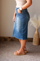 A person wearing a medium-wash blue denim midi skirt with a faded wash, paired with a beige top and brown platform slide sandals, standing on a textured gray carpet.