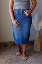 A person wearing a medium-wash blue denim pencil skirt with a front button closure and belt loops, paired with a gray top and turquoise sandals, standing indoors next to a woven vase with dried grass.