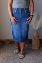 A person wearing a medium-wash blue denim pencil skirt with decorative silver embroidery on the front pockets, paired with a gray top and turquoise sandals, standing indoors next to a woven vase with dried grass.