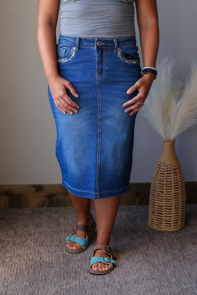 A person wearing a medium-wash blue denim pencil skirt with decorative silver embroidery on the front pockets, paired with a gray top and turquoise sandals, standing indoors next to a woven vase with dried grass.