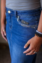 Close-up of a person wearing blue denim jeans with a decorative embroidered pocket and a black product tag tucked into the pocket.