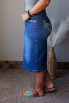 A person wearing a medium blue denim midi skirt with decorative silver embroidery on the back pocket, paired with brown slide sandals and beaded bracelets, standing in an indoor setting.