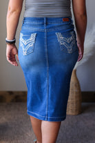 A person shown from the back wearing a fitted blue denim skirt with decorative white embroidery on the back pockets, paired with a gray top in an indoor setting.