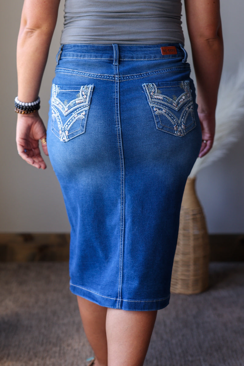 A person shown from the back wearing a fitted blue denim skirt with decorative white embroidery on the back pockets, paired with a gray top in an indoor setting.