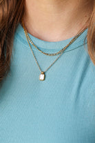 A close-up of a person wearing a layered necklace set, featuring a gold chain with a small rectangular gold pendant and a thicker gold chain, over a light blue ribbed top.