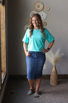 Woman in a turquoise shirt and blue skirt standing in a room with decorative wall art and a vase.