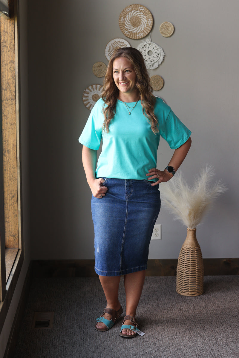 Woman in a turquoise shirt and blue skirt standing in a room with decorative wall art and a vase.