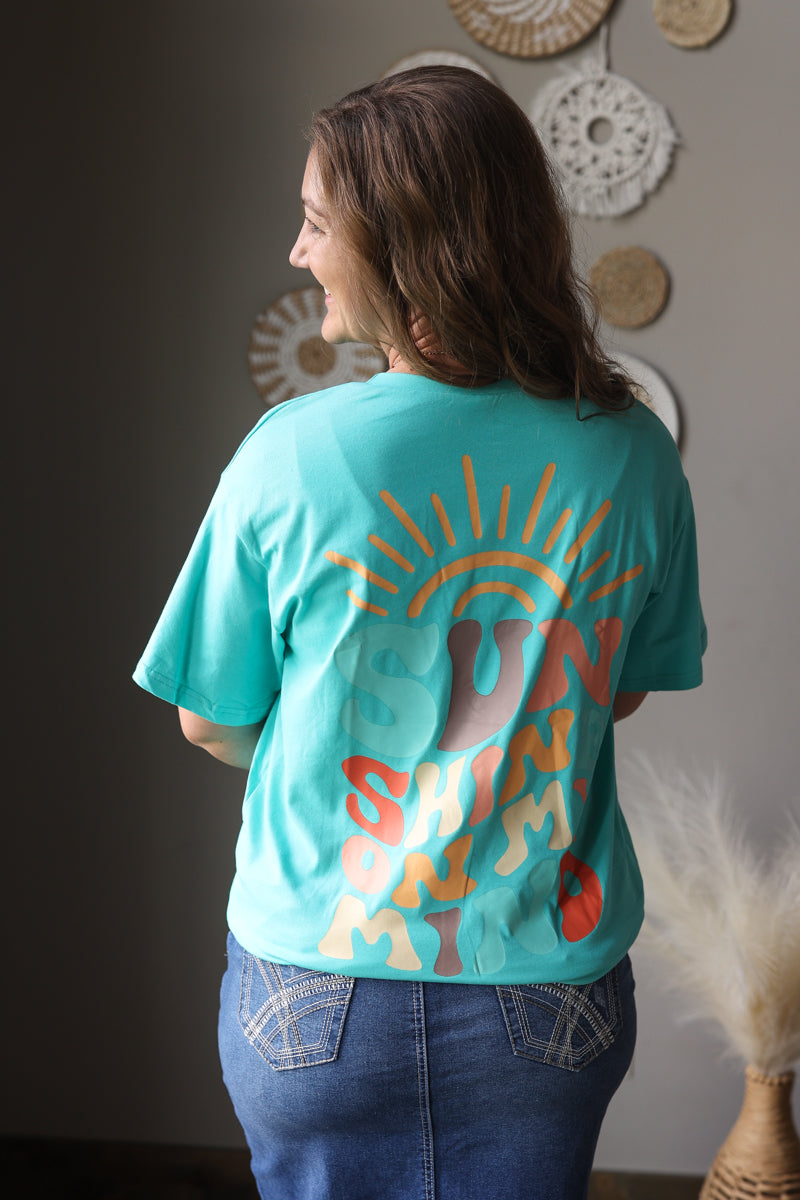 Person wearing a turquoise t-shirt with colorful designs and text, standing in a room with decorative items on the wall.