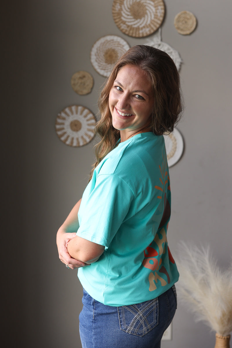 Woman wearing a teal shirt with a colorful design, standing against a gray wall with decorative plates.