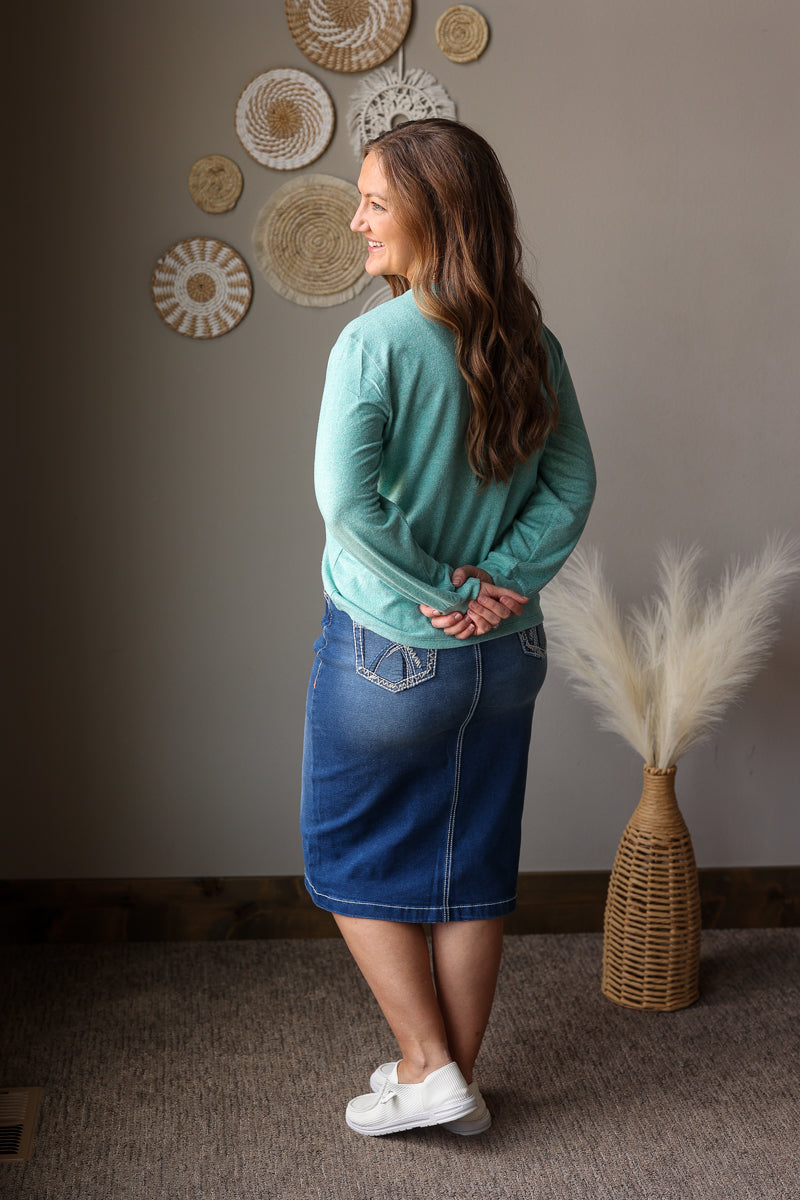 ash teal soft long sleeve top and denim skirt on a woman standing against a neutral wall for spring outfits