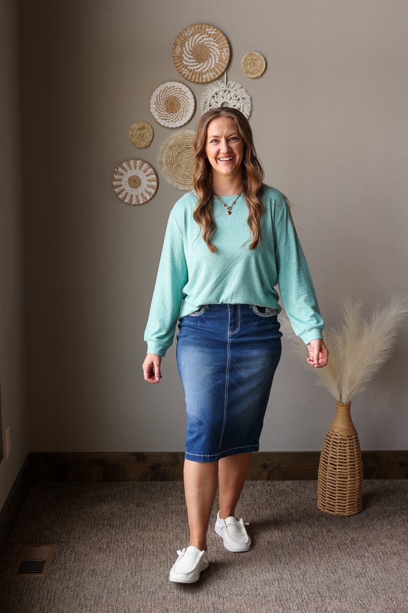 ash teal soft long sleeve top and denim skirt on a woman standing against a neutral wall for casual spring outfits