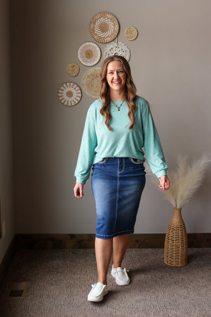 ash teal soft long sleeve top and denim skirt on a woman standing against a neutral wall for casual spring outfits