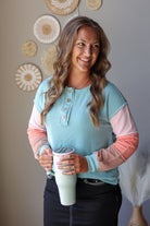 A woman smiling and holding a mint green tumbler, wearing a pastel color-blocked ribbed henley top with light blue, pink, and peach sections, set against a neutral wall with decorative sunburst wall hangings.