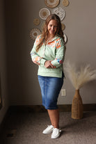 Woman wearing a light green hoodie with colorful sleeves and blue midi skirt standing in a room with decorative wall art.