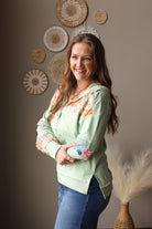 Woman wearing a light green blouse with floral cuffs and blue jean skirt, standing against a wall with decorative plates.