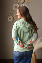 Woman wearing a light green top with colorful cuffs and floral patterns, standing against a neutral wall.