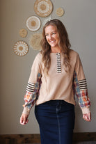 A woman wearing a beige long-sleeve top with striped and plaid patchwork sleeves, paired with a dark blue skirt, standing against a neutral wall with decorative woven wall art.