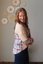 A woman wearing a color-blocked top with a beige knit body, a red and blue plaid panel, and striped sleeves, standing against a neutral wall with woven wall art.