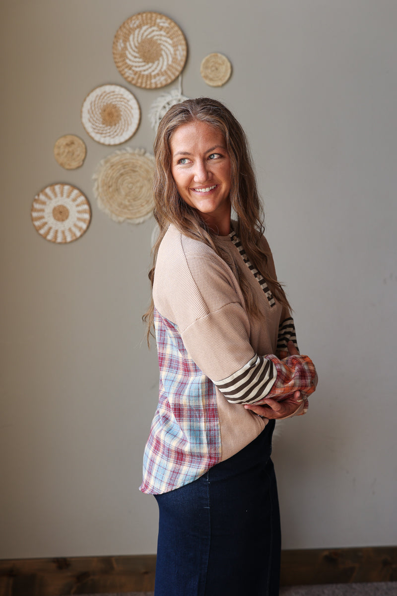 A woman wearing a beige and plaid button henley fall top, standing indoors against a neutral wall with woven wall decor.