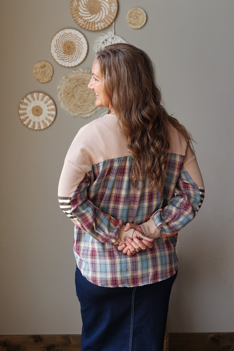 A woman with long brown hair is shown from behind wearing a color-blocked plaid top. The top features a beige yoke, tan sleeves, and a blue, red, and beige plaid body.