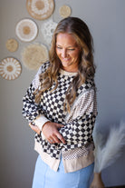 A woman wearing a bohemian-style patchwork jacket with black and white checkered panels, brown and beige gingham, and cream-colored trim, paired with light blue jean skirt. She is standing against a gray wall decorated with circular woven wall art.