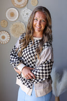 A smiling woman with long wavy light brown hair models a long-sleeve knit top featuring a black and white checkerboard pattern on the front and sleeves, with beige and brown checkered patterned panels on the sides and a beige hem. She is wearing light blue jean skirt against a gray wall decorated with circular woven wall art.