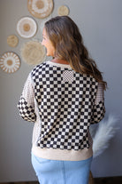 A woman shown from the back wearing a black and white checkerboard cropped sweater with beige ribbed trim, paired with light blue jean skirt, standing against a gray wall decorated with circular woven wall art.