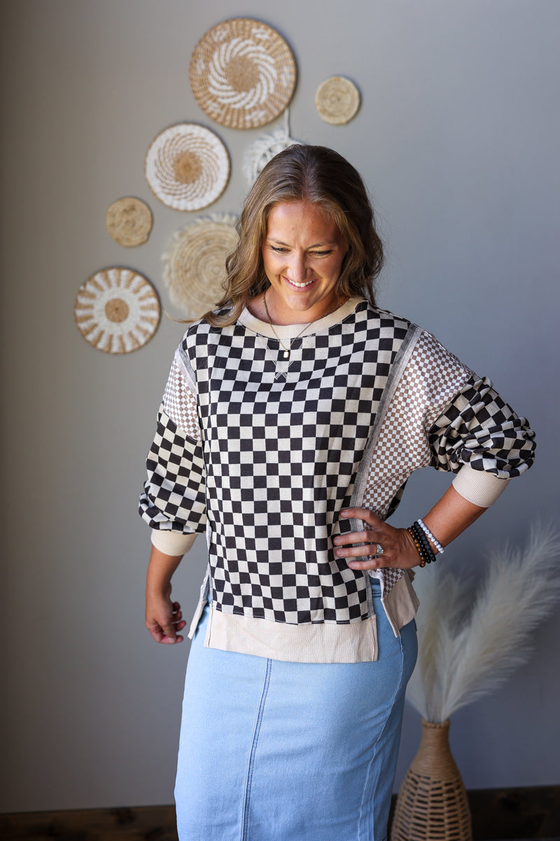 A woman wearing a black and white checkered knit sweater with beige mesh sleeves and a light blue denim skirt, standing in front of a gray wall decorated with circular woven wall art.