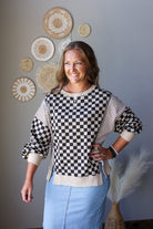 A woman modeling a black and white checkered long-sleeve top with beige trim and light blue skirt, standing against a gray wall decorated with circular woven wall art.