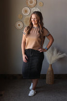 Woman posing in a room with decorative wall art and a vase. wearing a black denim skirt, white slip on shoes, and a brown animal print short sleeve top